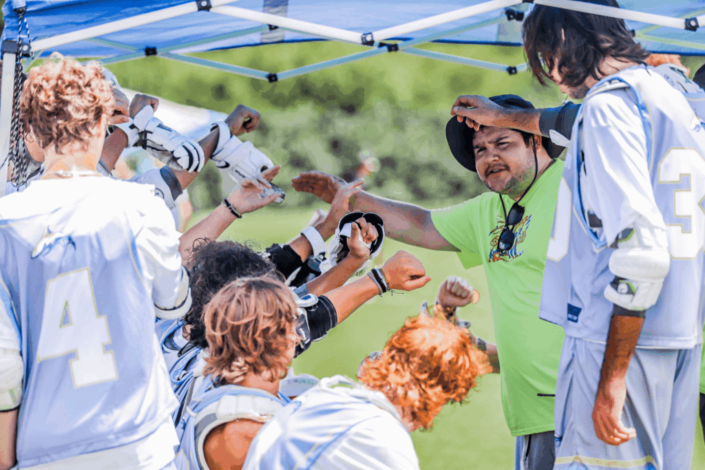 Group of lacrosse players huddled in with their coach, all raising their hands in solidarity