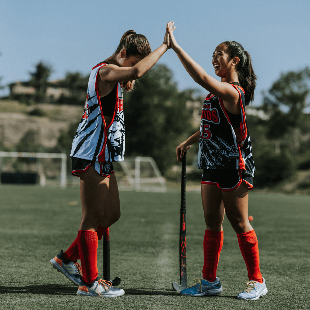 Two girls wearing full uniform and holding field hockey sticks high five each other while smiling