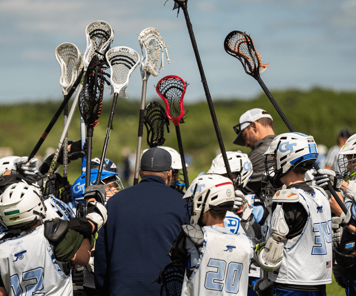 Coaches standing in a huddle with young lacrosse players who are holding up their sticks